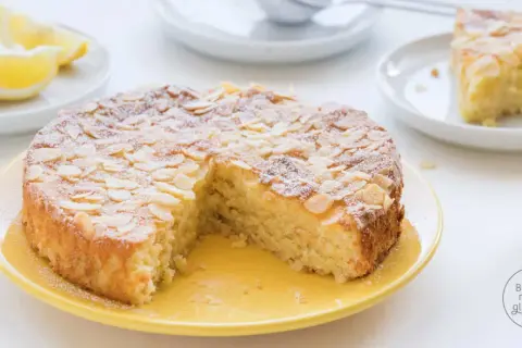 Saftiger Kuchen ohne Mehl, bestreut mit Mandelblättchen und Puderzucker. Dazu Zitronenspalten und eine ganze Zitrone.