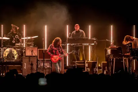 The War on Drugs band performs live, bathed in warm stage lights. A guitarist in a red plaid shirt plays center stage, with a drummer, keyboardist, and another musician on synths.