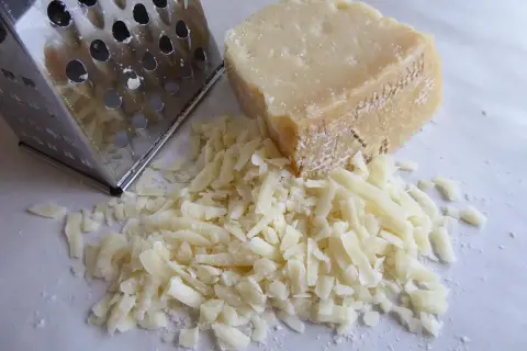 Un pezzo di formaggio a pasta cotta accanto a una grattugia, con formaggio grattugiato sparso.
