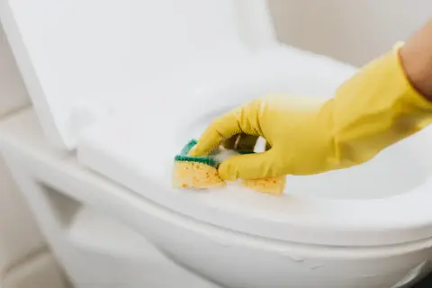 Ręka w żółtej rękawiczce czyści deskę sedesową gąbką. Dowiedz się, jak usunąć zapach chloru z pomieszczenia, by cieszyć się świeżością.