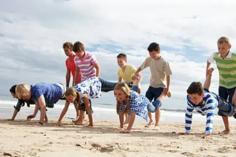 Niños compiten en juegos en la playa sin material, haciendo carreras de carretilla humana y otros desafíos divertidos.