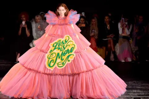 A model wears a voluminous pink tiered gown with a "Less is More" sign, embodying a bold camp aesthetic on the runway.