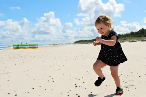 Radosne wakacje z dziećmi w Polsce. Mała dziewczynka biega po piaszczystej plaży, ciesząc się słońcem i chmurami na niebie.