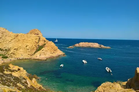 En solig dag vid Ile Rousse. Turkost vatten, klippor och båtar i bukten. En segelbåt syns i fjärran.