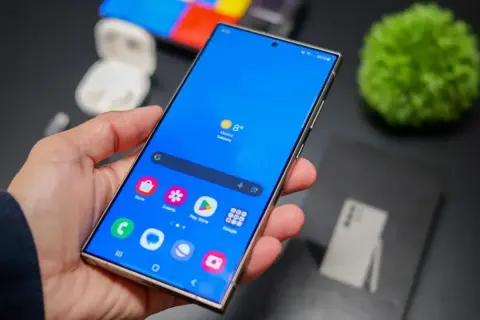Mano sujetando un smartphone con pantalla azul vibrante, mostrando iconos de aplicaciones y el tiempo. La interfaz, **que es One UI**, se ve fluida y organizada.