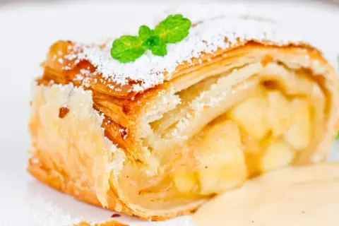 Ein Stück Apfelstrudel mit Puderzucker und Minze, serviert mit Vanillesauce. Perfekt für ein Apfelstrudel Rezept!