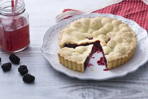 Une délicieuse tarte aux mûres, avec une part coupée, sur une assiette ornée. Des mûres fraîches et un verre de jus rouge complètent la scène.