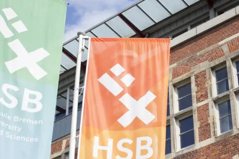 Zwei Fahnen der Hochschule Bremen City University of Applied Sciences wehen vor einem Backsteingebäude.
