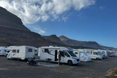 Un grupo de autocaravanas low cost aparcadas en un terreno pedregoso, con montañas y cielo azul de fondo.