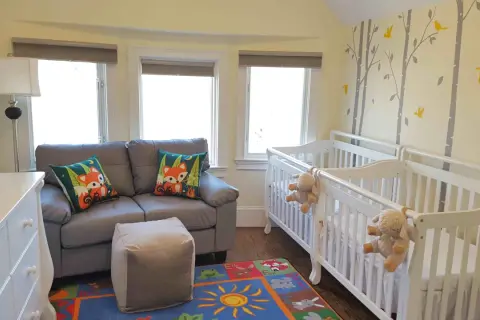 A charming nursery with two white cribs, perfect for nursery sets for twins. A grey sofa with fox-themed pillows and a colorful rug complete the cozy space.