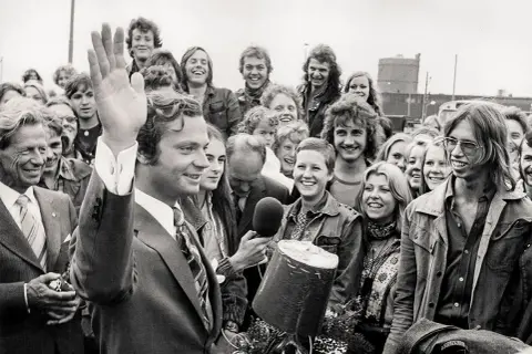 Kung Carl XVI Gustaf vinkar glatt till en folkmassa. En ung kvinna håller fram en mikrofon, kanske för att intervjua en av kungens barn eller någon annan i den glada gruppen.