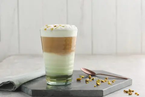 Un latte pistache aux couches distinctes, couronné de mousse et de pistaches concassées. Une cuillère en cuivre repose à côté.
