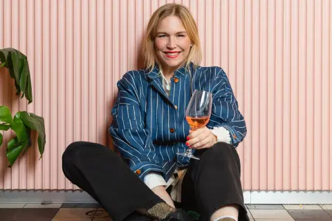 Frida Lund blogg: En glad kvinna med ett glas rosévin sitter på golvet framför en rosa vägg.