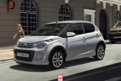 Srebrny Citroën C1 z czarnymi nakładkami na błotniki przemierza miejską ulicę.