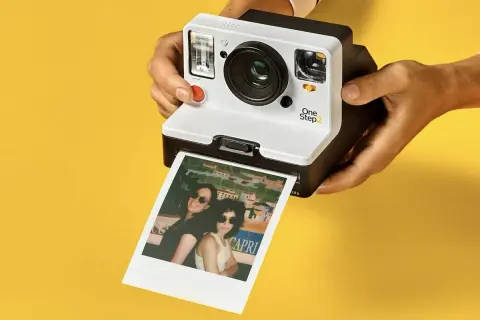 Aparat Polaroid One Step 2 drukuje zdjęcie dwóch kobiet w okularach przeciwsłonecznych na tle kolorowego krajobrazu.