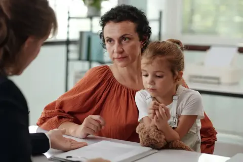 Matka z córką słuchają diagnozy autyzmu. Dziecko tuli misia, a kobieta patrzy z uwagą.
