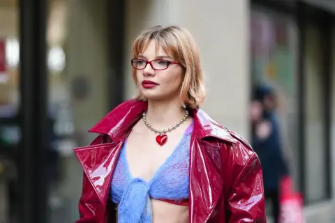 Frau mit roten Brillen und einem stufigen Bob trägt eine glänzende rote Jacke über einem blauen Strickoberteil mit Herz-Halskette.