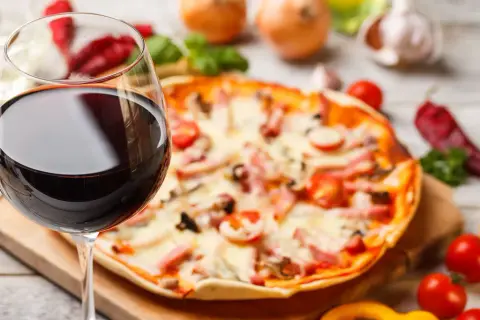Ein Glas Rotwein neben einer Pizza mit Tomaten und Schinken. Die perfekte Kombination: welcher Wein zu Pizza?
