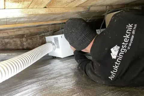 Installering av ventilation i friggebod. En tekniker arbetar med en vit ventilationsenhet under träbjälkar.