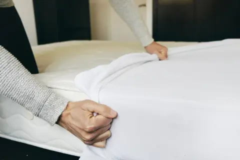 Hands pulling a white mattress pad over a mattress. This is part of how to clean a mattress pad.