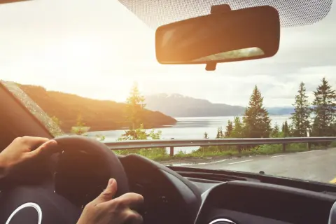 Hände am Lenkrad, Blick durch die Windschutzscheibe auf eine bergige Landschaft mit See. Die Sonne scheint, und man fragt sich, ob man durch das Glas braun wird.