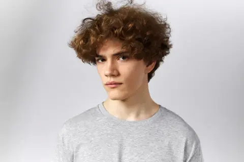 Joven con un peinado rizos hombre, cabello castaño y rizado, viste camiseta gris.