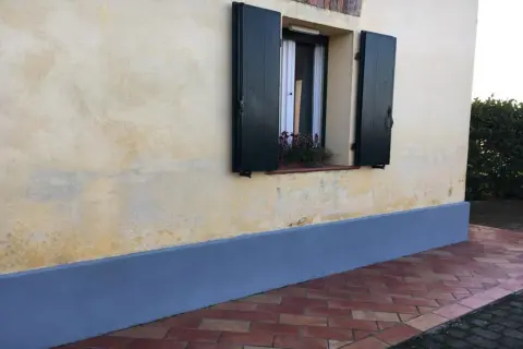 Muro con rinzaffo storico, una finestra con persiane verdi e un davanzale fiorito. Pavimentazione in cotto.