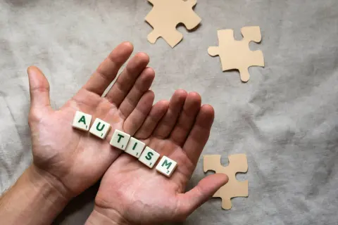 Twee handen houden de letters vast die 'AUTISME' spellen. Puzzelstukjes liggen verspreid, symbolisch voor het spectrum.
