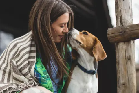 Ein Hund sucht die Aufmerksamkeit seiner Besitzerin. Sie kuscheln eng aneinander, umgeben von rustikalen Holzelementen.