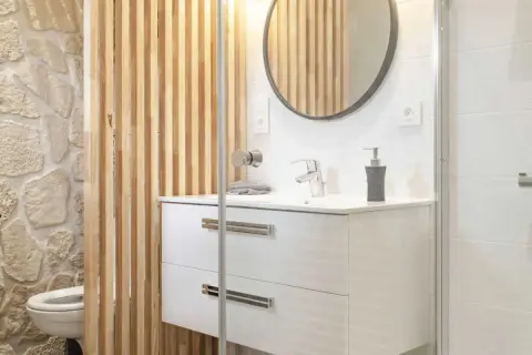 Salle de bain moderne avec un lavabo suspendu, un miroir rond et une **installation WC** discrète derrière un paravent en bois.