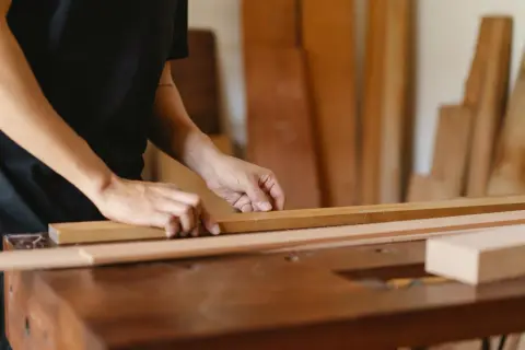 Manos midiendo una tabla de madera en un taller. Se aprecian otras tablas en el fondo.