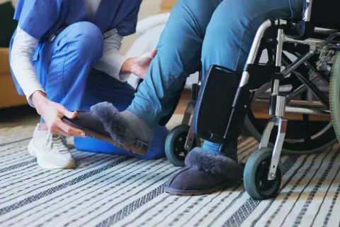 Opiekunka zakłada kapcie osobie na wózku inwalidzkim.