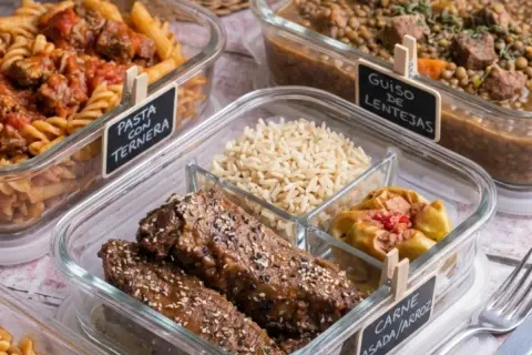 Carne asada con arroz, pasta con ternera y guiso de lentejas en recipientes de vidrio, listos para llevar.