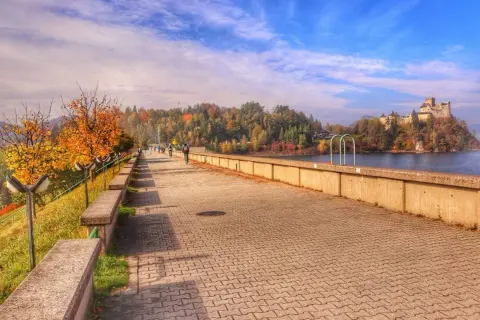 Jesioro Czorsztyńskie jesienią. Spacerowicze i rowerzyści na promenadzie, w tle zamek i kolorowe drzewa.