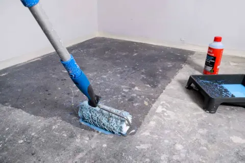 Rodillo aplicando líquido para quitar sintasol del suelo. Bandeja con pintura azul y botella de producto al lado.