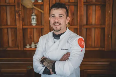Carlos Maldonado, chef con tatuajes y barba, sonríe con los brazos cruzados frente a una estantería de madera.
