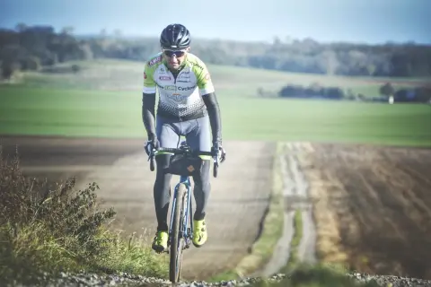 Kolarz w stroju sportowym jedzie rowerem po szutrowej drodze wśród pól.