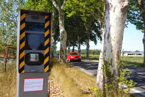 Panneau de **détecteur de radar interdit** sur le bord de la route, avec une voiture rouge passant à proximité.