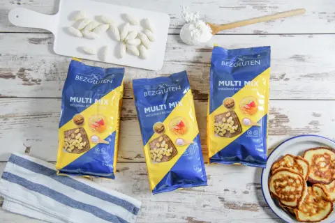 Trzy opakowania mieszanki bezglutenowej Multi Mix, placuszki, mąka i białe paluszki na desce.