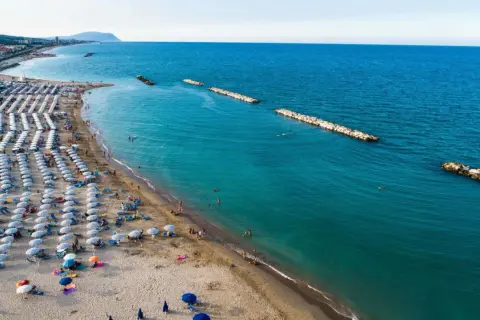Ampia spiaggia libera a Civitanova Marche, con file ordinate di ombrelloni e lettini, bagnanti che si godono il mare turchese e dighe frangiflutti in pietra.