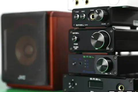 Stosik czarnych urządzeń audio SMSL, w tym SMSL M6 Mini DAC, obok brązowej kolumny głośnikowej.