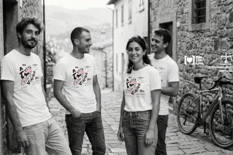 Quattro amici sorridono in un vicolo di pietra, indossando magliette con un disegno di bici e la scritta "A tutta virra". Un'espressione del vocabolario dialetto marchigiano che evoca passione.