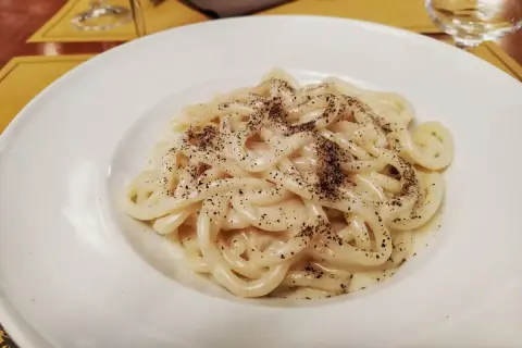 Piatto di pici cacio e pepe, pasta fresca condita con pecorino romano e pepe nero macinato fresco, servita su un piatto bianco.