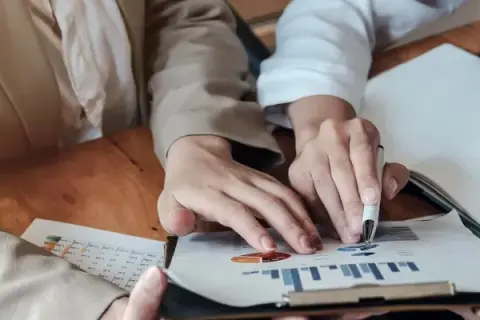 Dłonie analizujące wykresy i tabelę na drewnianym stole.