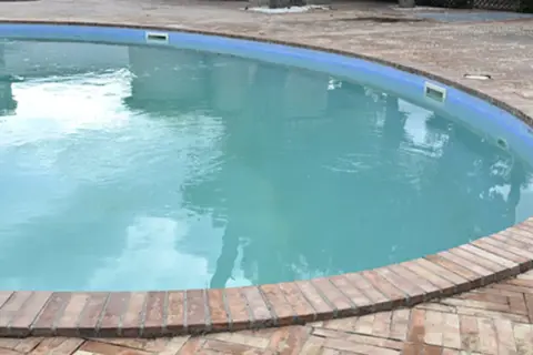 Agua de piscina blanquecina, con reflejos del cielo y rodeada por un borde de ladrillos.
