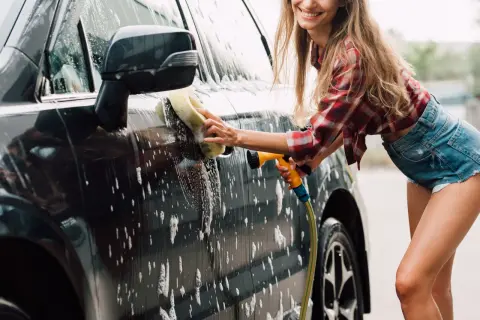 Uśmiechnięta kobieta myje auto gąbką i wężem. Domowe sposoby na czysty samochód to świetna zabawa!