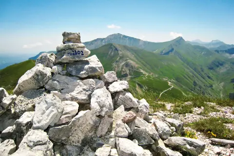 Cairn con M. 2102, simbolo di un'avventura di trekking nei Monti Sibillini. Panoramica mozzafiato sulle verdi valli e sentieri tortuosi.