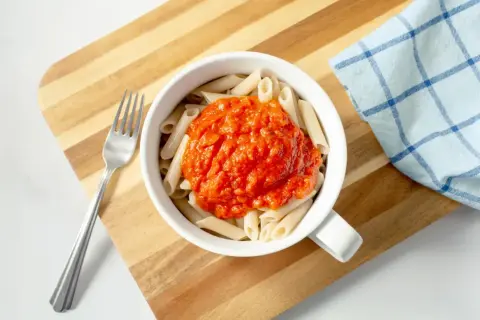 Makaron penne z sosem pomidorowym w białej misce, obok widelec i ściereczka.