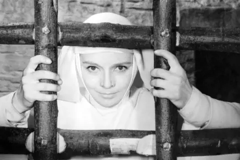 Matka Joanna od Aniołów, w białym habicie, trzyma się drewnianego krzyża. Jej oczy patrzą wprost na widza.