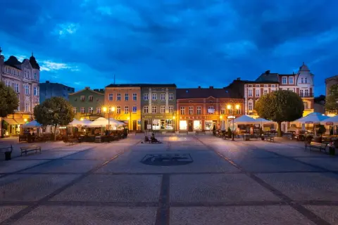 Wieczorny rynek w Kościerzynie, z oświetlonymi kamienicami i ogródkami restauracyjnymi.